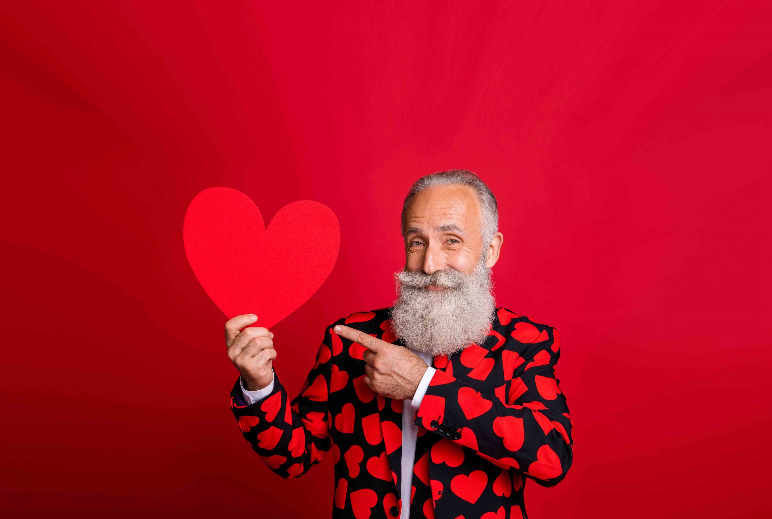 Photo of funky mature man cupid amour hold big paper heart shape indicating finger creative postcard wear stylish hearts pattern suit blazer shirt tie isolated red color background
