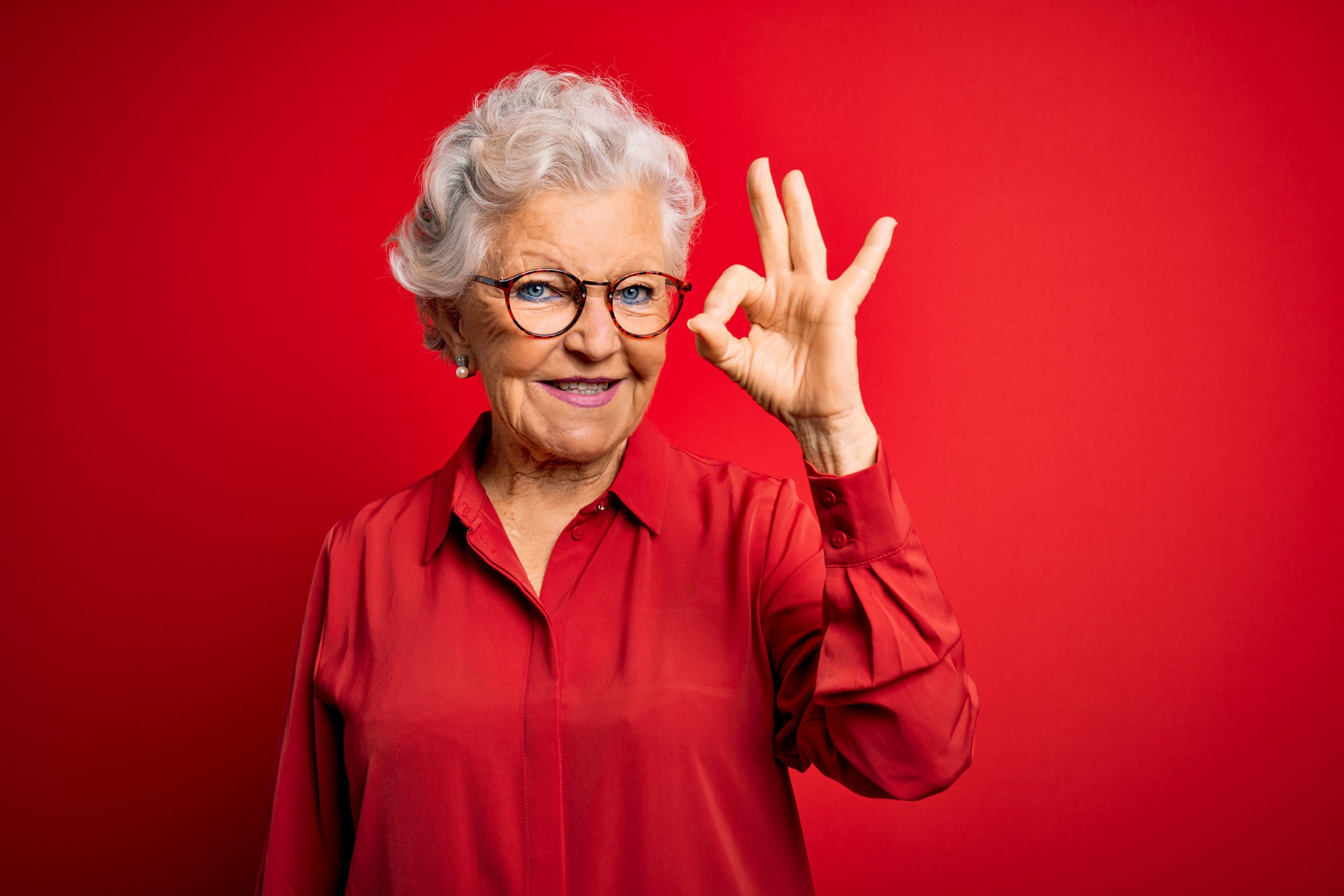 Senior beautiful grey-haired woman wearing casual shirt and glasses over red background smiling positive doing ok sign with hand and fingers. Successful expression.